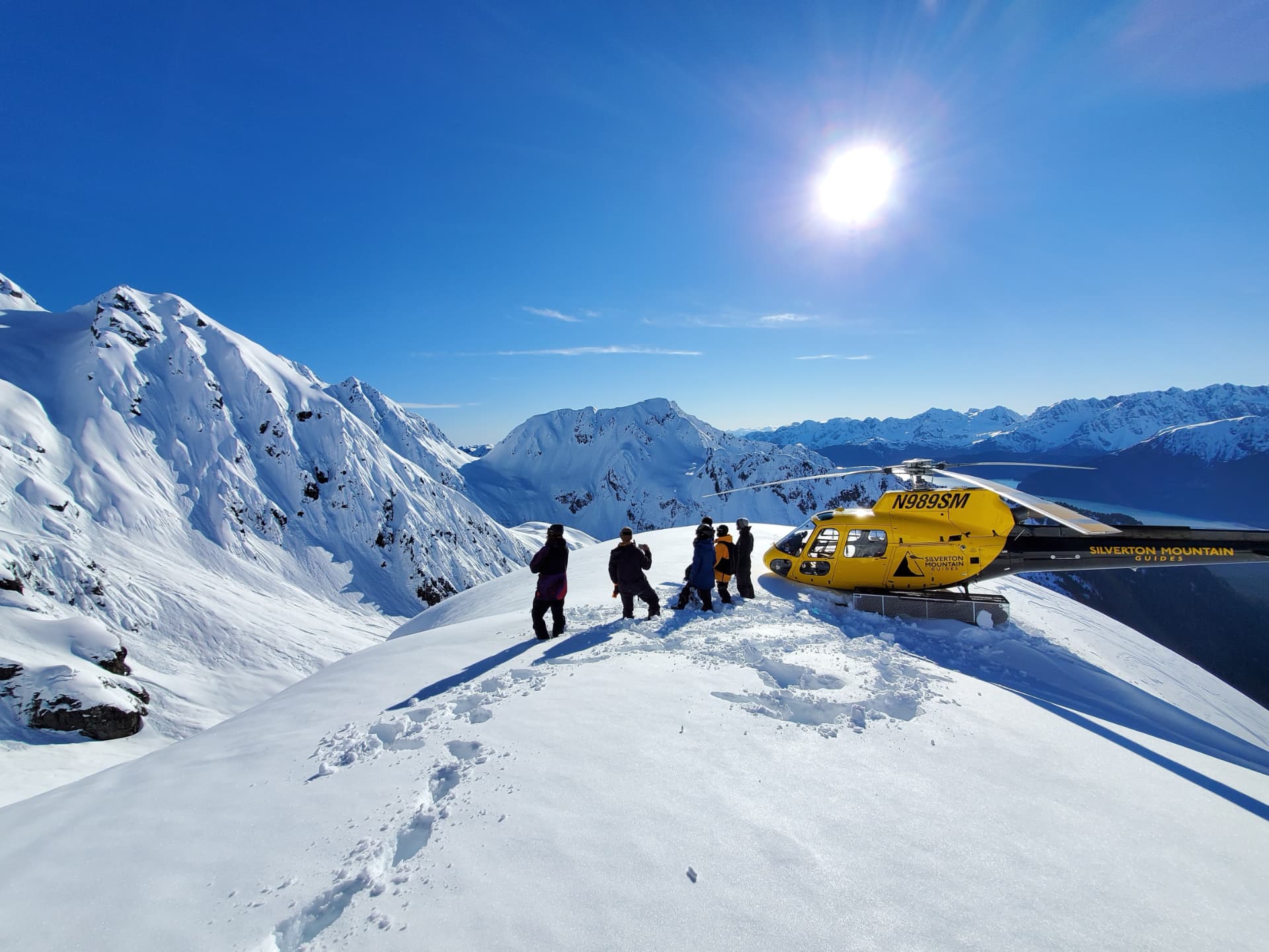 SMG skiers and yellow helicopter on a snowy Seward ridge under a bright sun