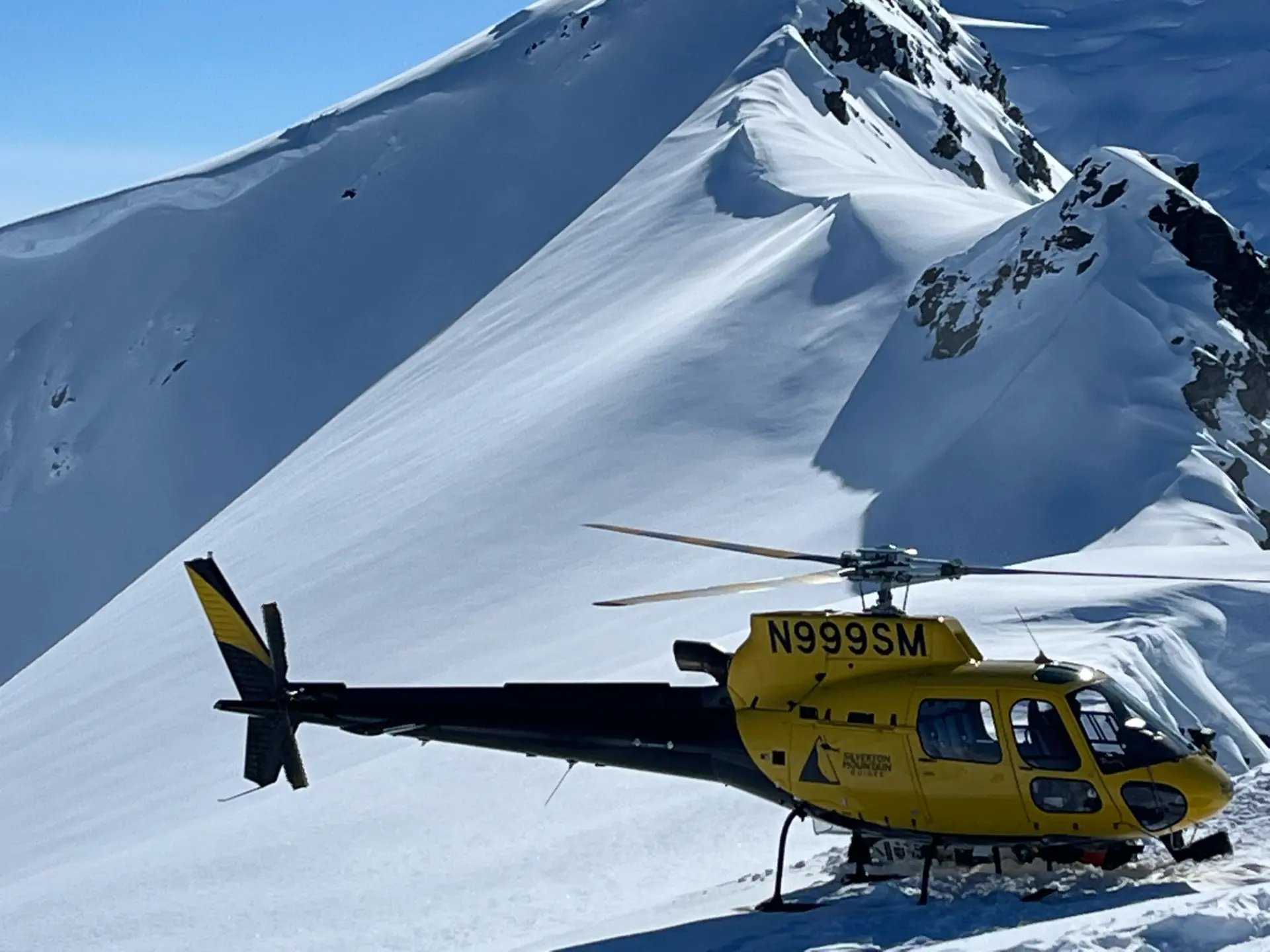 SMG yellow helicopter N999SM on a snowy Alaskan ridge with massive peaks behind