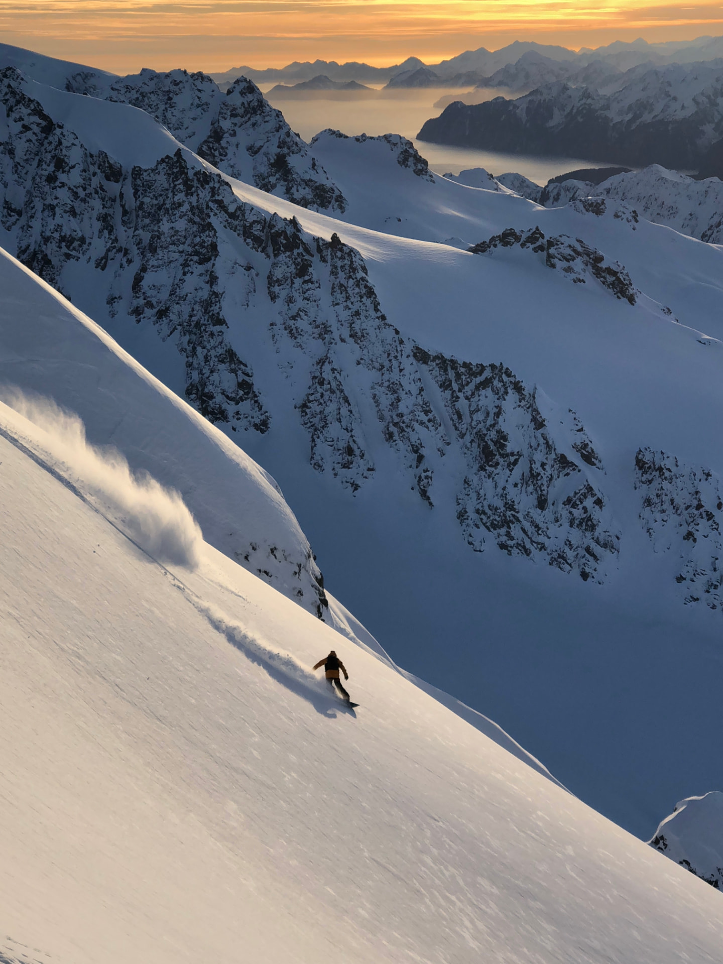 A snowboarder descends a steep, untouched Alaskan powder face at sunset with endless mountain ranges behind